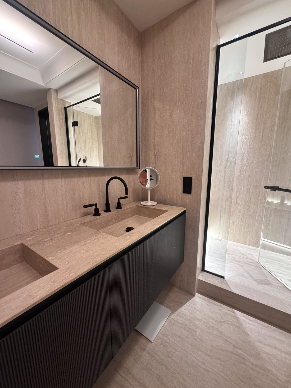 Salle de bains du duplex Maubourg : travertin au sol et aux murs, meuble double vasque noir et douche vitrée